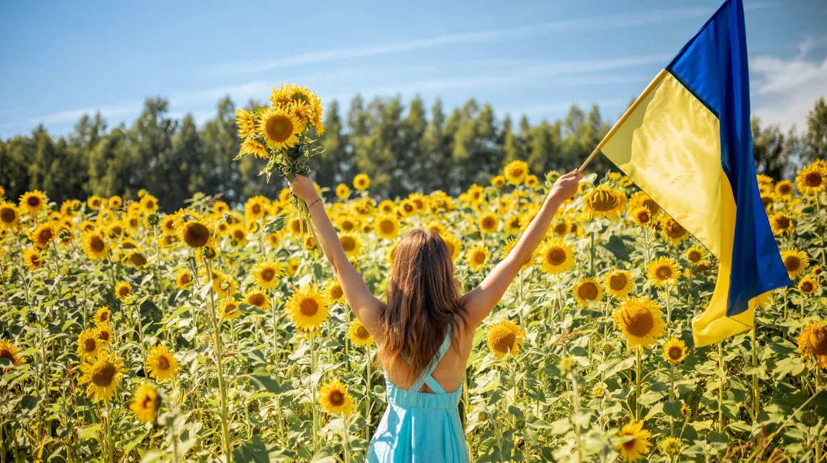 Kobieta stojąca tyłem na polu słoneczników, trzymająca flagę Ukrainy i bukiet słoneczników w rękach